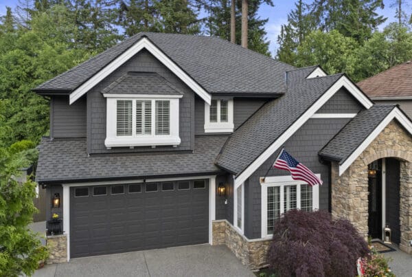 Two-story gray house with white trim, stone accents, and an American flag by the front entrance. Surrounded by trees and greenery, the house has a double garage and manicured landscaping.