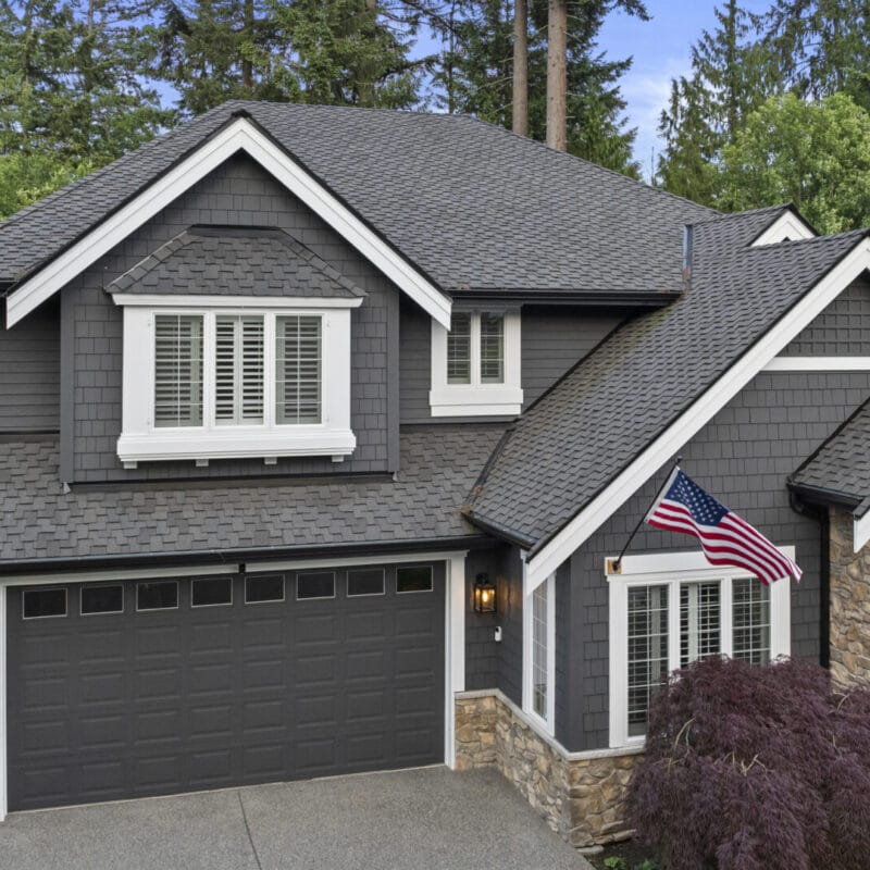 Two-story gray house with white trim, stone accents, and an American flag by the front entrance. Surrounded by trees and greenery, the house has a double garage and manicured landscaping.