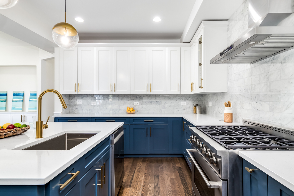Modern kitchen with white upper cabinets, blue lower cabinets, white countertops, stainless steel appliances, gold hardware, a large gas stove, a farmhouse sink, and pendant lighting over a wood floor.