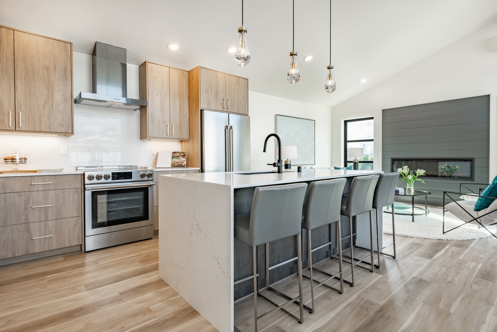 Modern kitchen with light wood cabinets, stainless steel appliances, a large island with four gray barstools, pendant lights overhead, and an open living area with a fireplace and large windows.