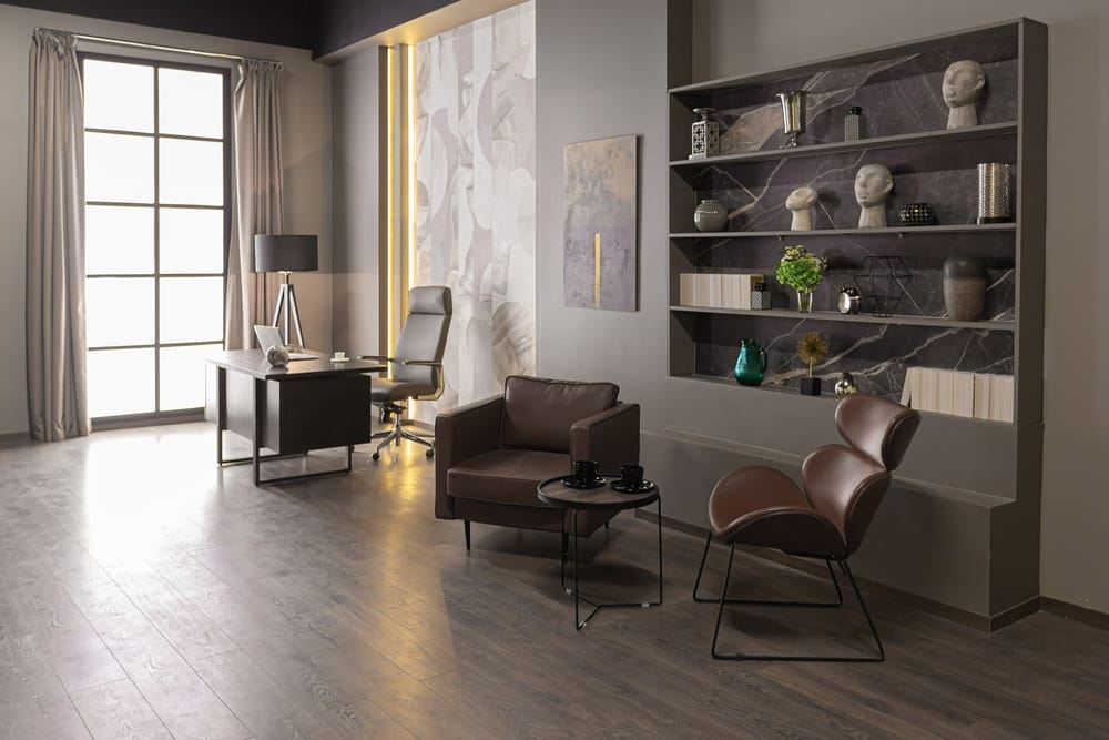 Modern office with a desk, lamp, and chair by large windows. There are two contemporary armchairs with small tables, built-in shelves with books, decor, and sculptures. Neutral tones and natural light create a stylish atmosphere.