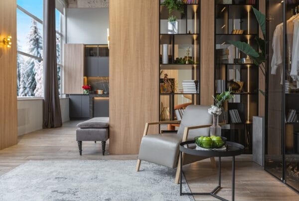 Modern living space with light wood floors, a gray armchair, round black table with green apples, open shelving, and large windows showing a snowy outdoor scene. A kitchen and gray bench are visible in the background.