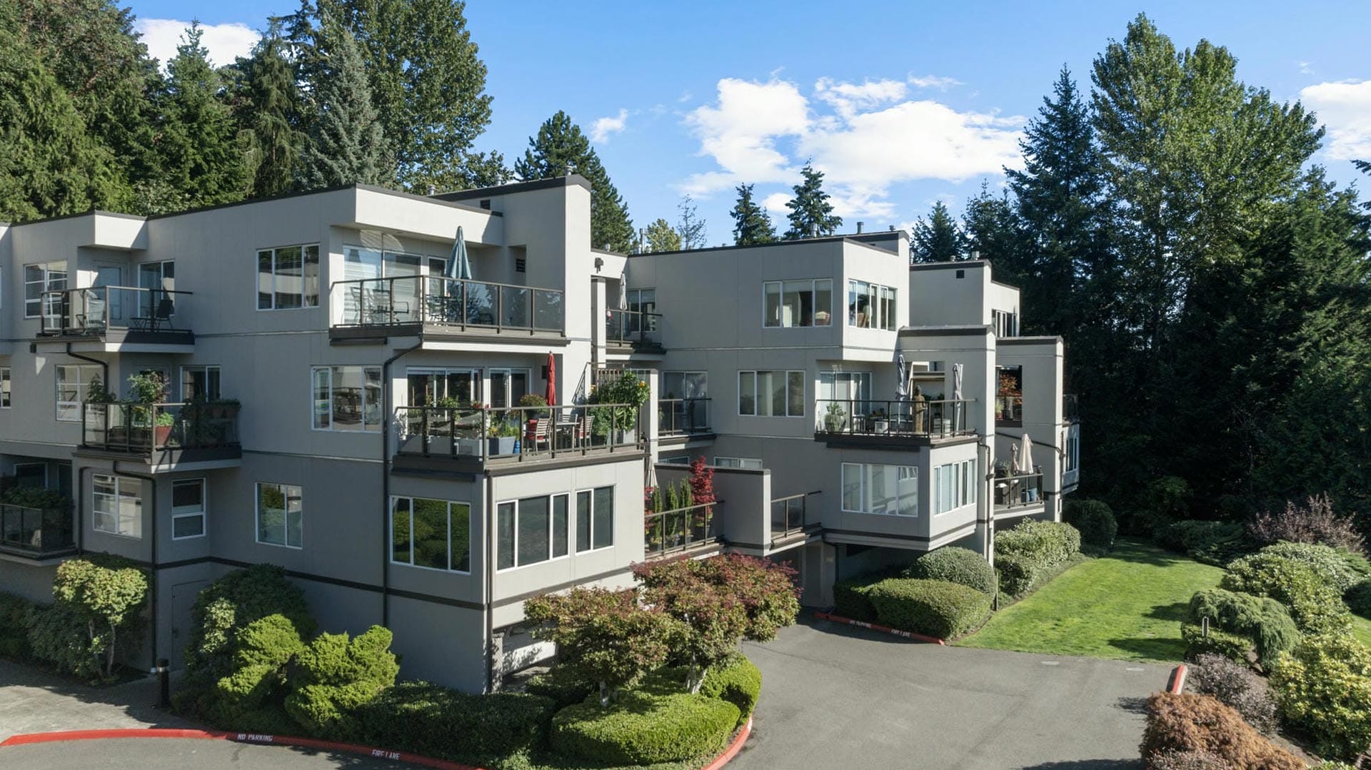 Modern gray apartment building with multiple balconies, surrounded by lush green trees and well-maintained shrubs, under a blue sky with scattered clouds.