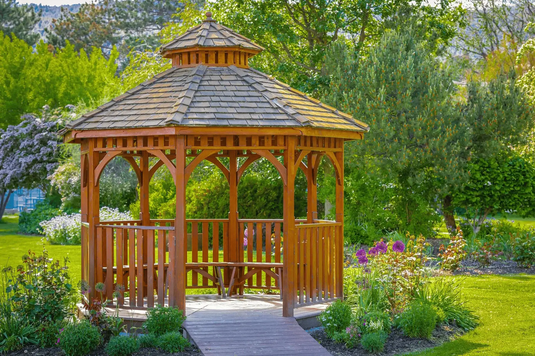A wooden gazebo with a shingled roof stands in a lush garden, surrounded by green grass, blooming flowers, and trees under a bright, sunny sky.