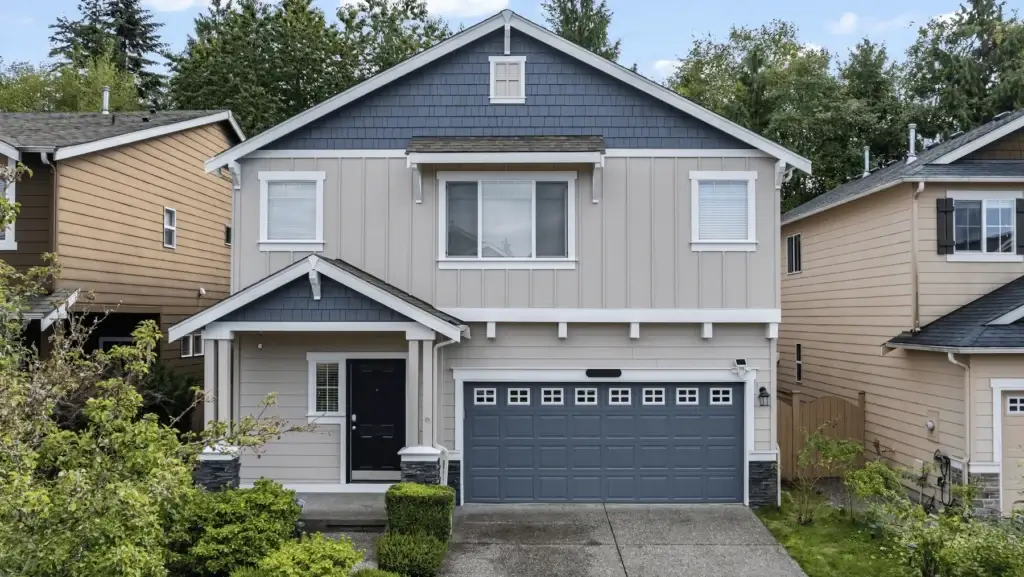 Two-story suburban house with gray siding, dark blue accents, a double garage, black front door, small front yard, and trees in the background. Neighboring houses are visible on both sides.
