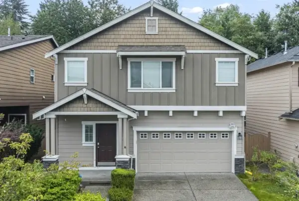 Two-story suburban house with taupe siding, white trim, a double garage, and a covered front porch. The house is surrounded by greenery and neighboring homes, with a concrete driveway and walkway.