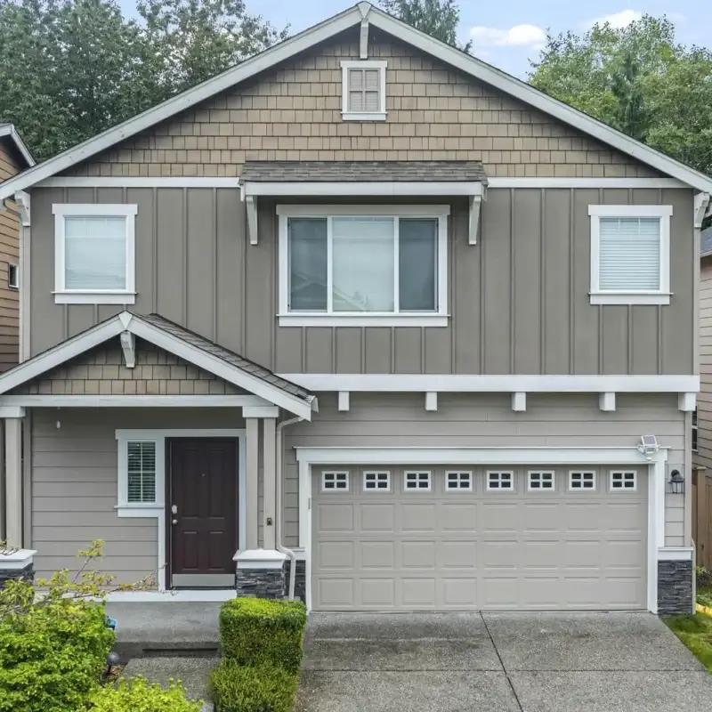 Two-story suburban house with taupe siding, white trim, a double garage, and a covered front porch. The house is surrounded by greenery and neighboring homes, with a concrete driveway and walkway.