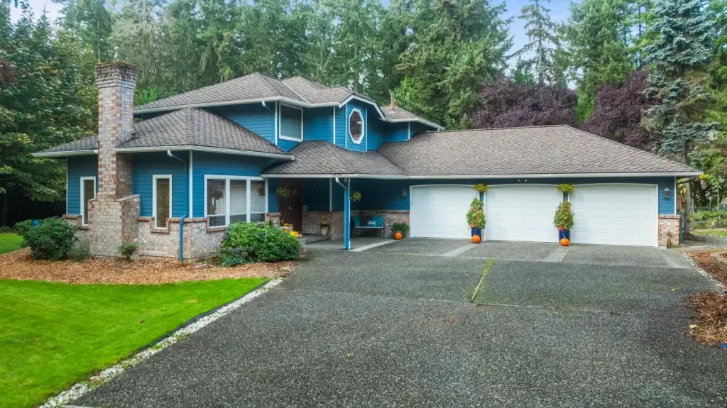 A two-story blue house with white trim, a large attached three-car garage, brick chimney, well-kept lawn, and trees in the background. Hanging plants and flower pots decorate the entrance and driveway.