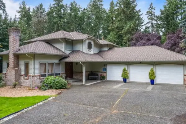 Two-story suburban house with a large driveway, three-car garage, brick accents, covered porch, and surrounded by tall trees and lush greenery.
