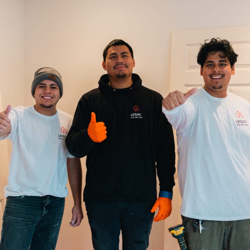 Three men stand indoors in front of white doors, smiling and giving thumbs up. Two wear white shirts with a company logo and one wears a black hoodie and orange gloves. The room appears to be under renovation.