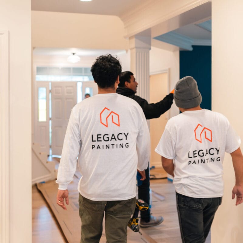 Two people wearing “Legacy Painting” shirts walk through a doorway inside a house, following another person who is pointing toward a room. The space appears to be under renovation, with bare walls and minimal furnishings.