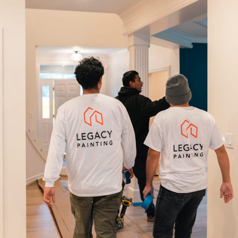 Two workers wearing Legacy Painting shirts walk through a house interior with painting tools, following another person who is pointing ahead. The space is bright with hardwood floors and white walls.