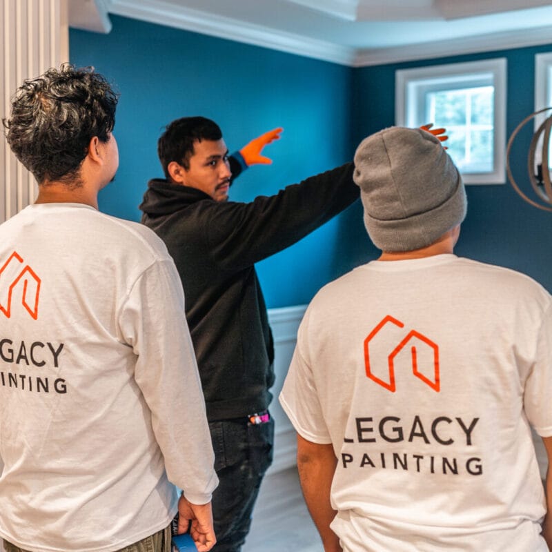 Three people stand in a room with blue walls and white trim; one person in a black hoodie gestures toward the windows, while two others in Legacy Painting shirts watch and listen attentively.