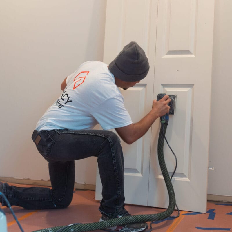 A person in a beanie and white t-shirt kneels on the floor while sanding a white door with an electric sander in a room under renovation. Blue painters tape marks the floor and baseboards.