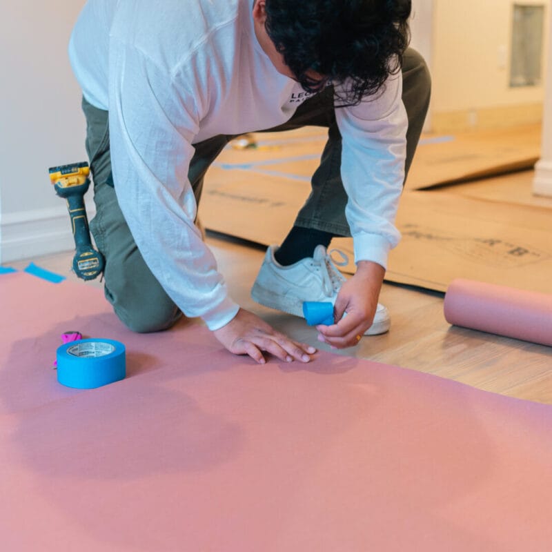 A person kneels on a wooden floor, securing a pink protective sheet with blue painters tape. Nearby are a roll of the same sheet and more tape, with cardboard sheets in the background.