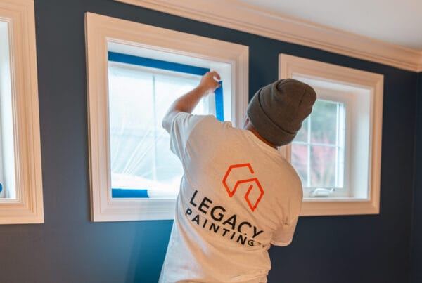 A person wearing a gray beanie and a Legacy Painting shirt applies blue painters tape around a window in a room with dark blue walls and white trim.