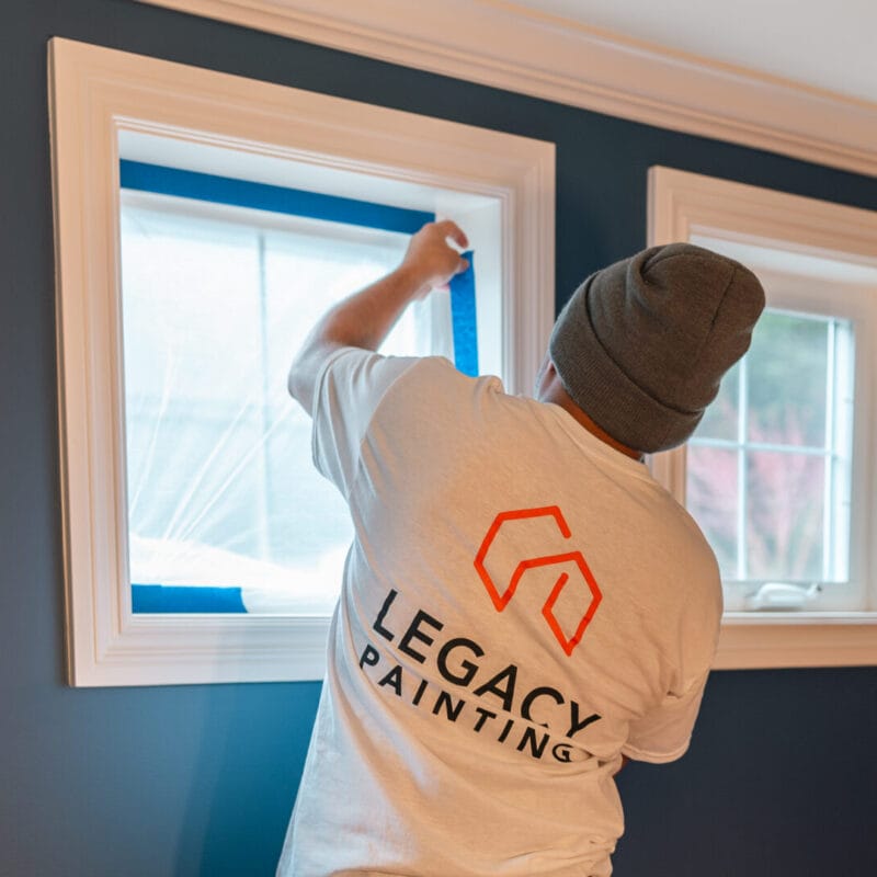 A person wearing a gray beanie and a Legacy Painting shirt applies blue painters tape around a window in a room with dark blue walls and white trim.