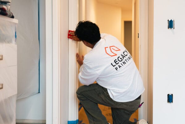 A person wearing a LEGACY PAINTING shirt kneels while using a sanding block on a doorway frame in a home under renovation. The area is prepped with plastic sheets and painter’s tape.
