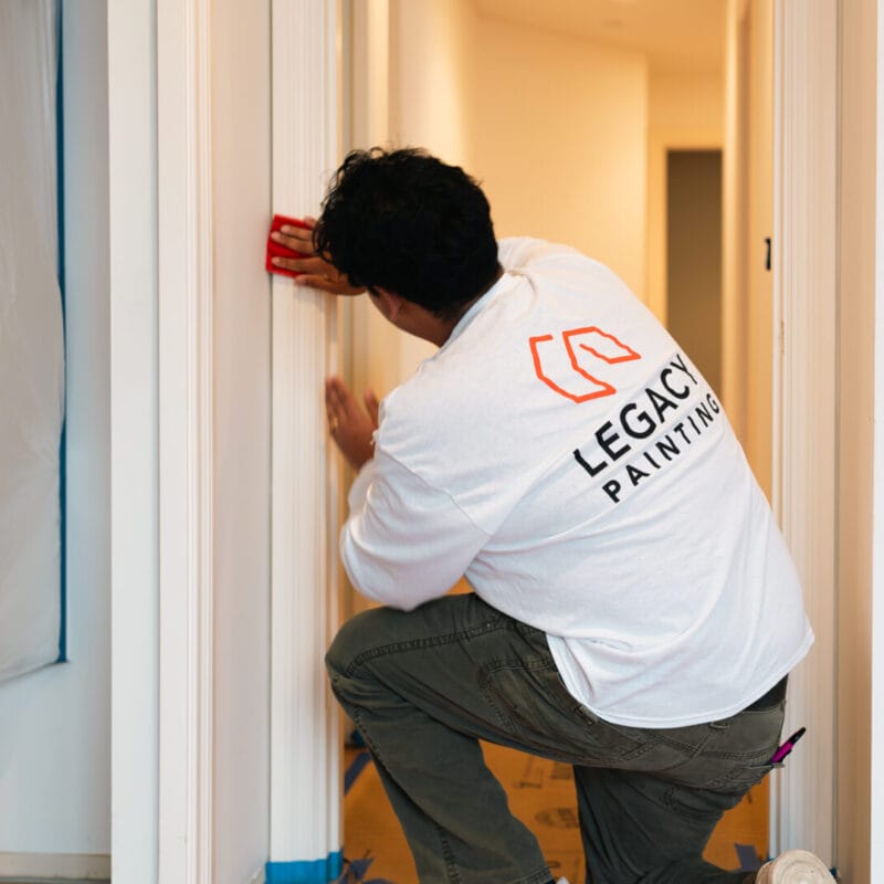 A person wearing a LEGACY PAINTING shirt kneels while using a sanding block on a doorway frame in a home under renovation. The area is prepped with plastic sheets and painter’s tape.