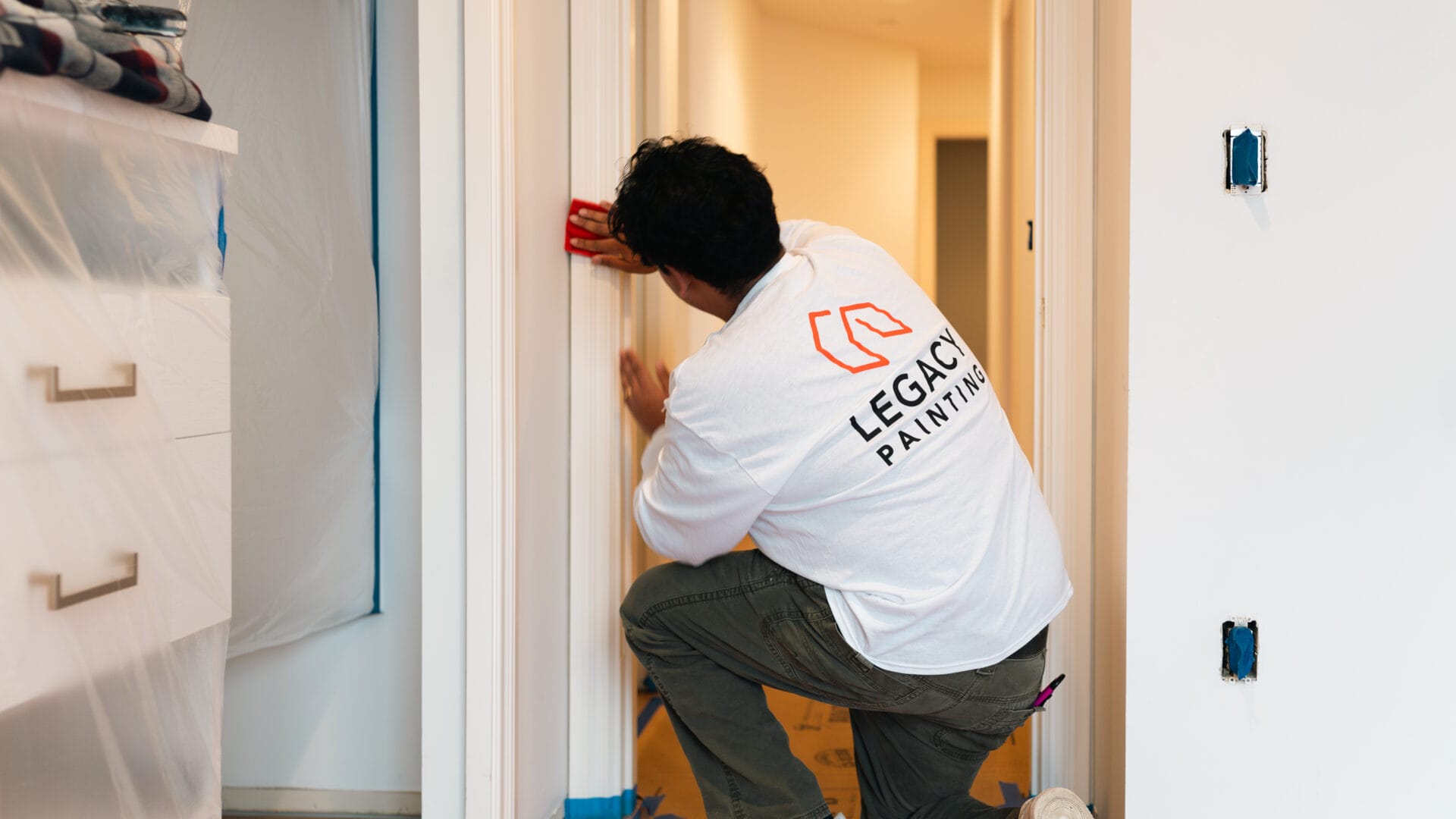 A person wearing a LEGACY PAINTING shirt kneels while using a sanding block on a doorway frame in a home under renovation. The area is prepped with plastic sheets and painter’s tape.