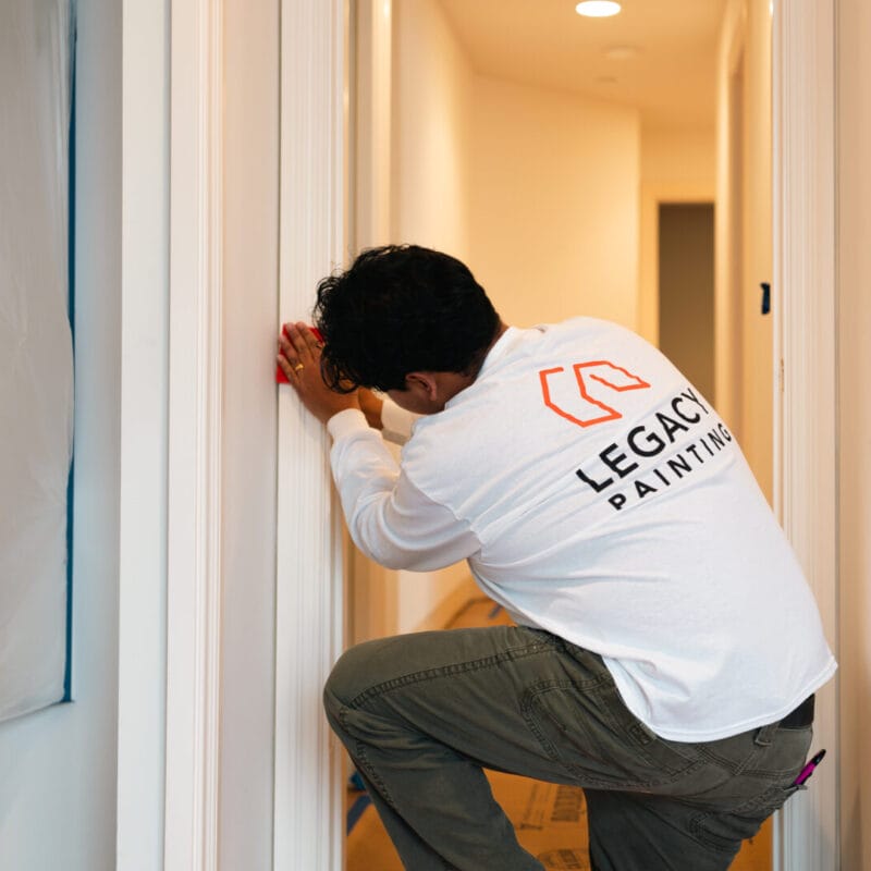 A person wearing a Legacy Painting shirt kneels in a hallway, working on a door frame in a home under renovation, with unpainted electrical outlets visible on the walls.
