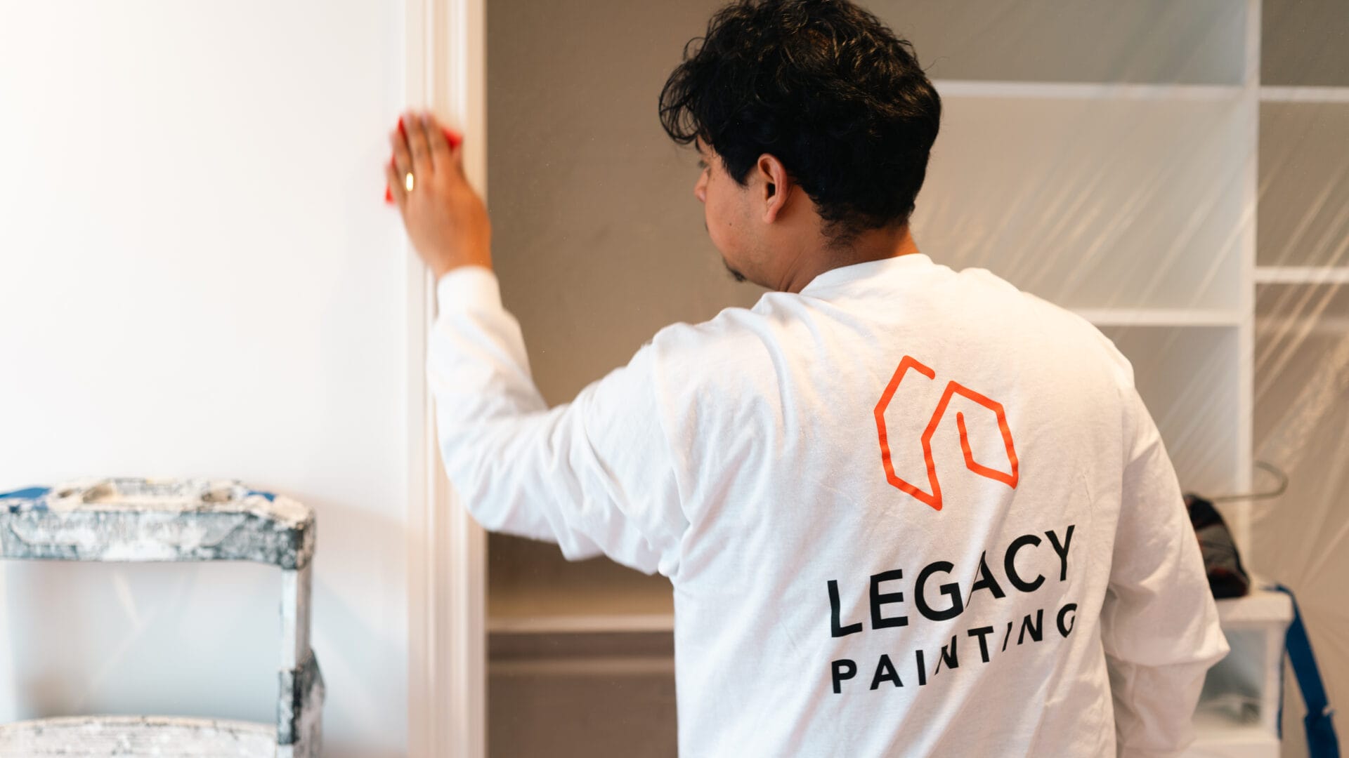 A person wearing a white “Legacy Painting” shirt sands a doorframe. Shelving and a step ladder are visible in the background.