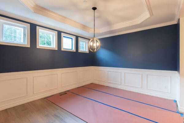 A room with blue upper walls, white wainscoting, four rectangular windows, a modern hanging light fixture, and paper covering part of the wooden floor, likely for renovation or painting.