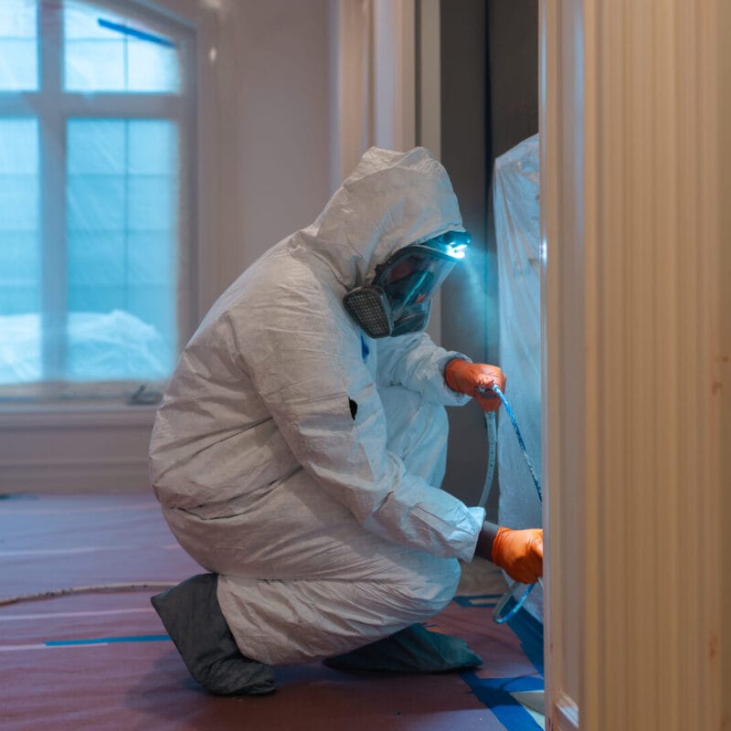 A person in full protective gear, including a hooded suit, gloves, and a respirator mask, kneels indoors while taping plastic sheeting to a doorway during a containment or cleaning process.
