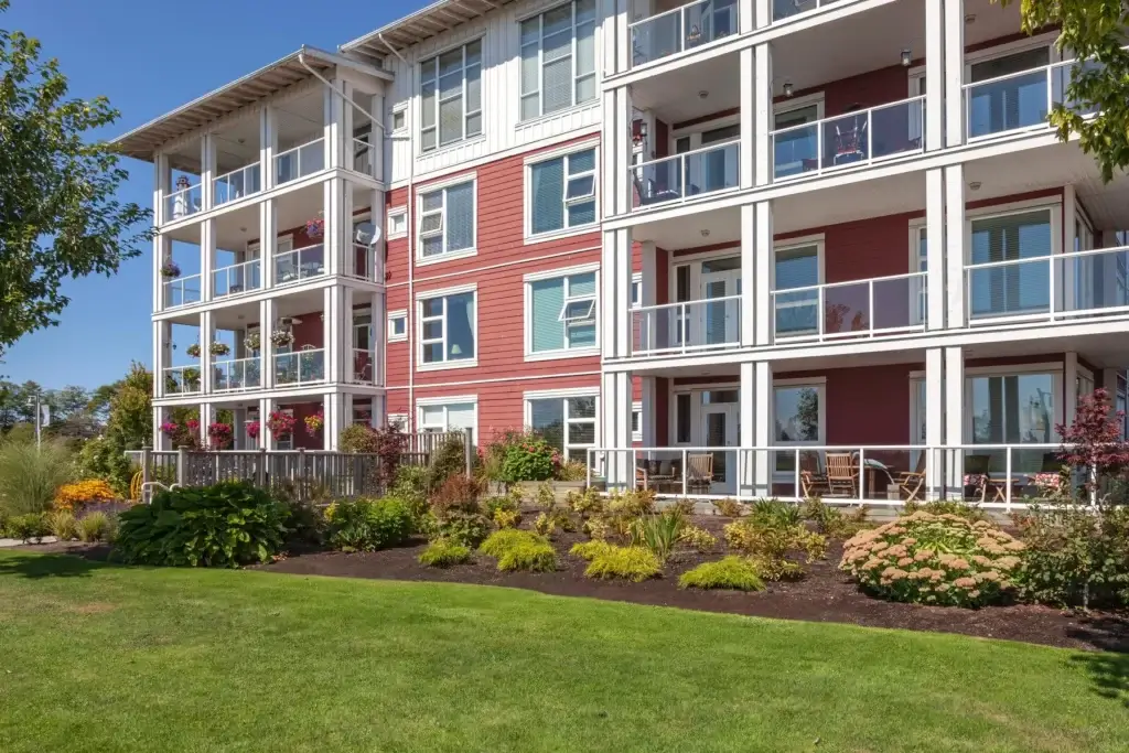 A modern, multi-story apartment building with red siding, white trim, and glass balcony railings. The landscaped yard features green grass, shrubs, and small trees under a clear blue sky.