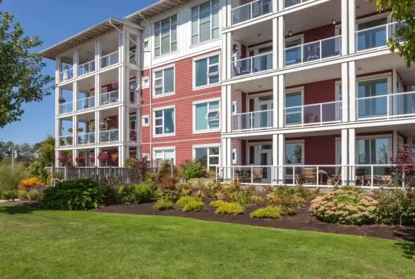 A modern, multi-story apartment building with red siding, white trim, and glass balcony railings. The landscaped yard features green grass, shrubs, and small trees under a clear blue sky.